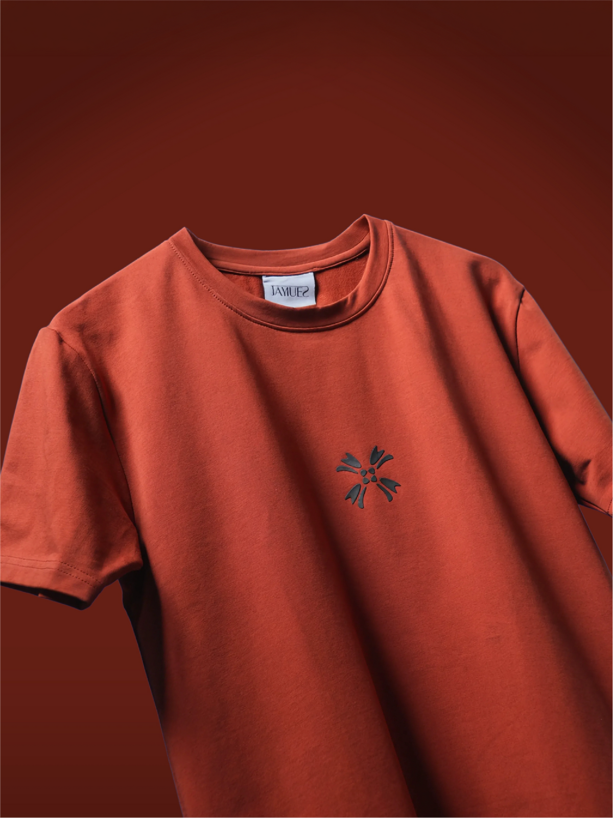 A rust colored t-shirt with a small dark black hd printed logo mark on the chest, displayed against a matching dark reddish-brown background.